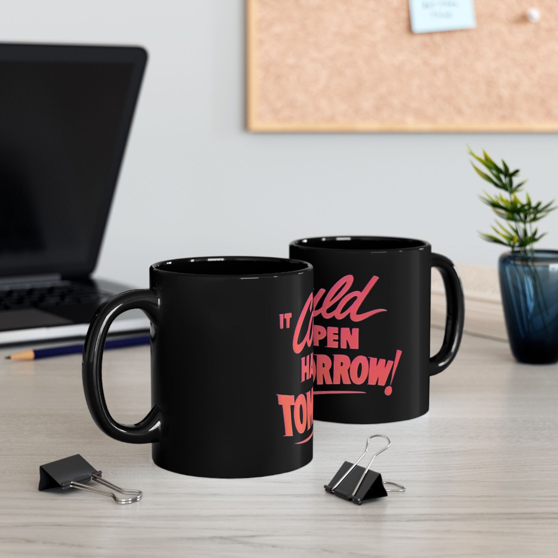 Two black mugs with pink text on a desk with office supplies and a plant.