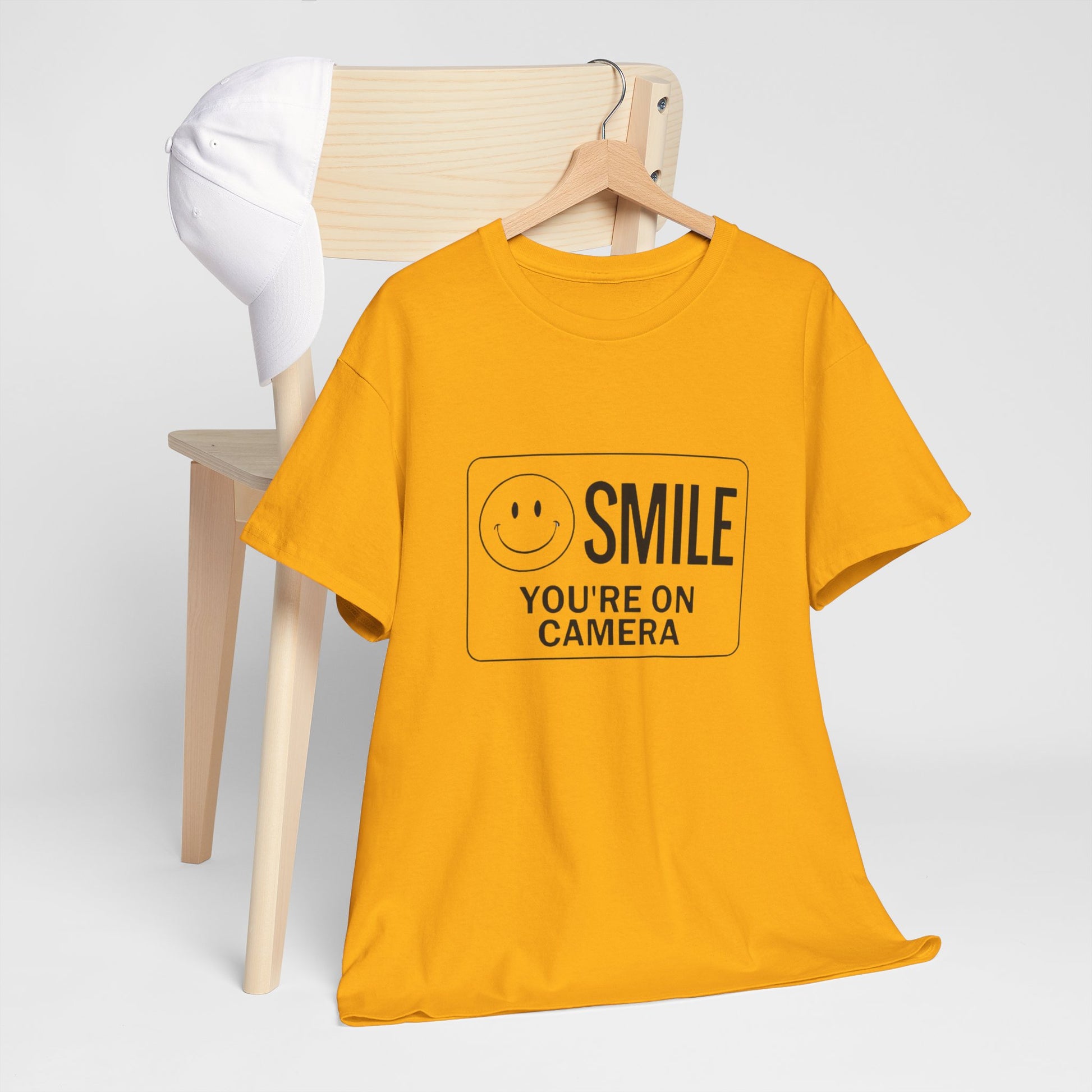 Yellow t-shirt with 'SMILE YOU'RE ON CAMERA' text and smiley face on a hanger against a white background